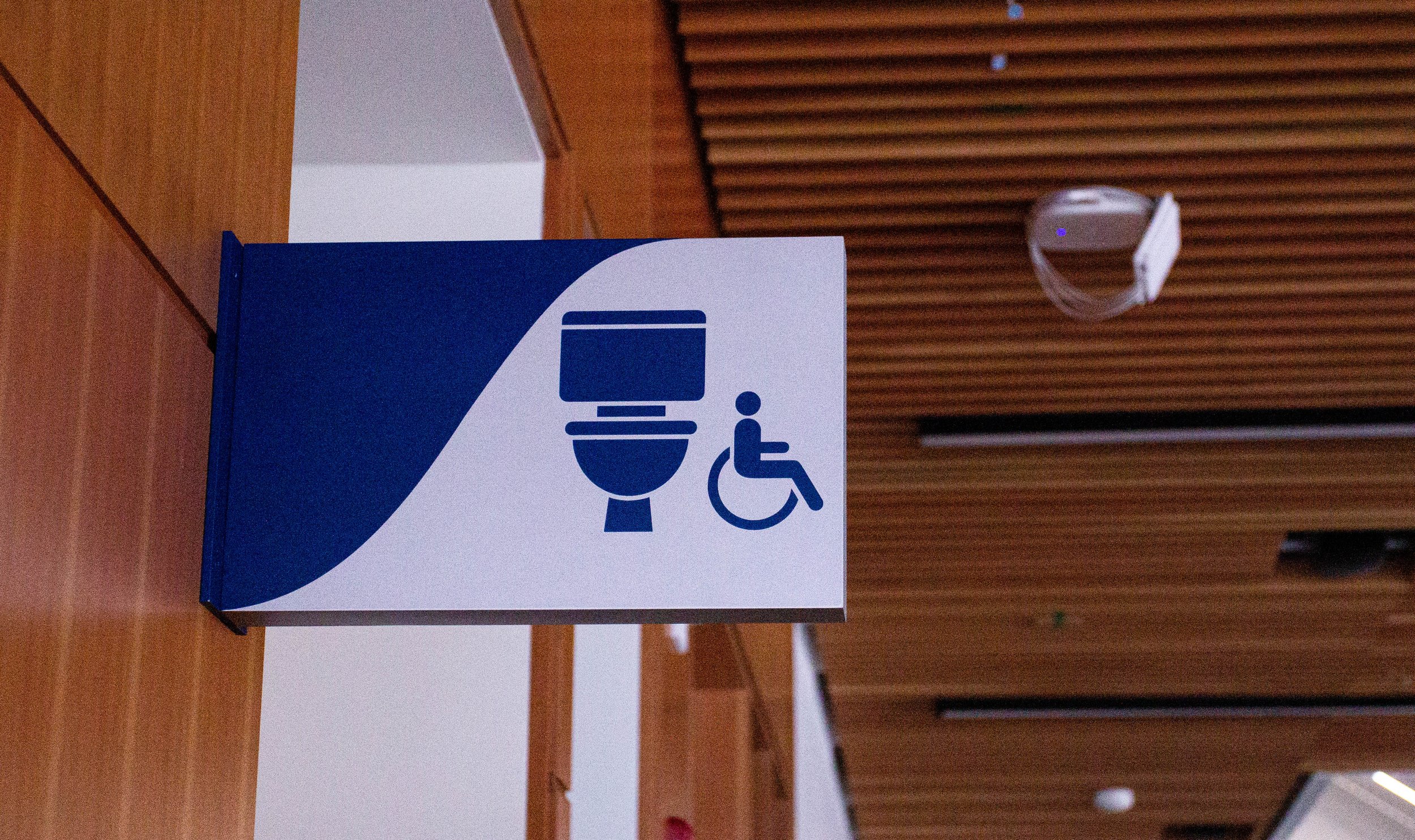 A view of a wooden panelled wall with a two-toned washroom sign: one side is a navy background, on the right a toilet icon next to the accessibility symbol.