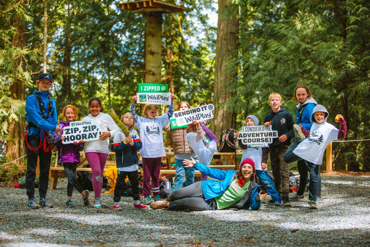 A group of cheerful children and instructors at WildPlay Victoria, holding signs with phrases like 'ZIP, ZIP, HOORAY' and 'I CONQUERED THE ADVENTURE COURSE!' during a week-long adventure camp.